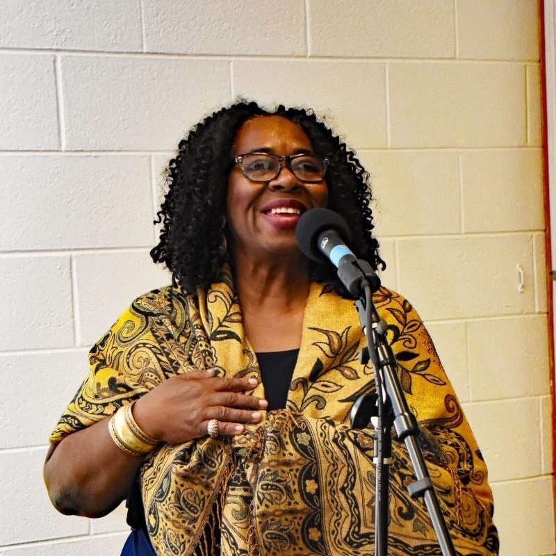 A Black woman stand at mic with her hand on her chest, smiling.